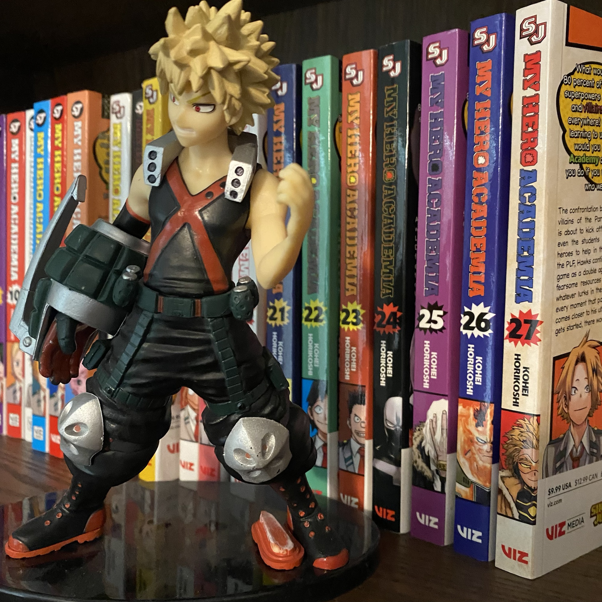 A collection of the My Hero Academia books up to volume 27 with a figurine of Bakugo in front.